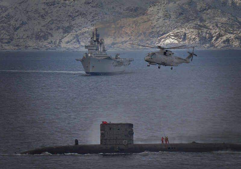HMS Talent conducts a high line transfer with a Merlin helicopter as HMS Ark Royal passes in the background